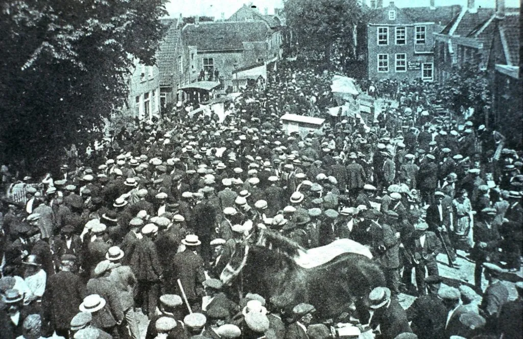 Oude zwart-witfoto van een straat vol mensen met platte petten tijdens de paardenmarkt